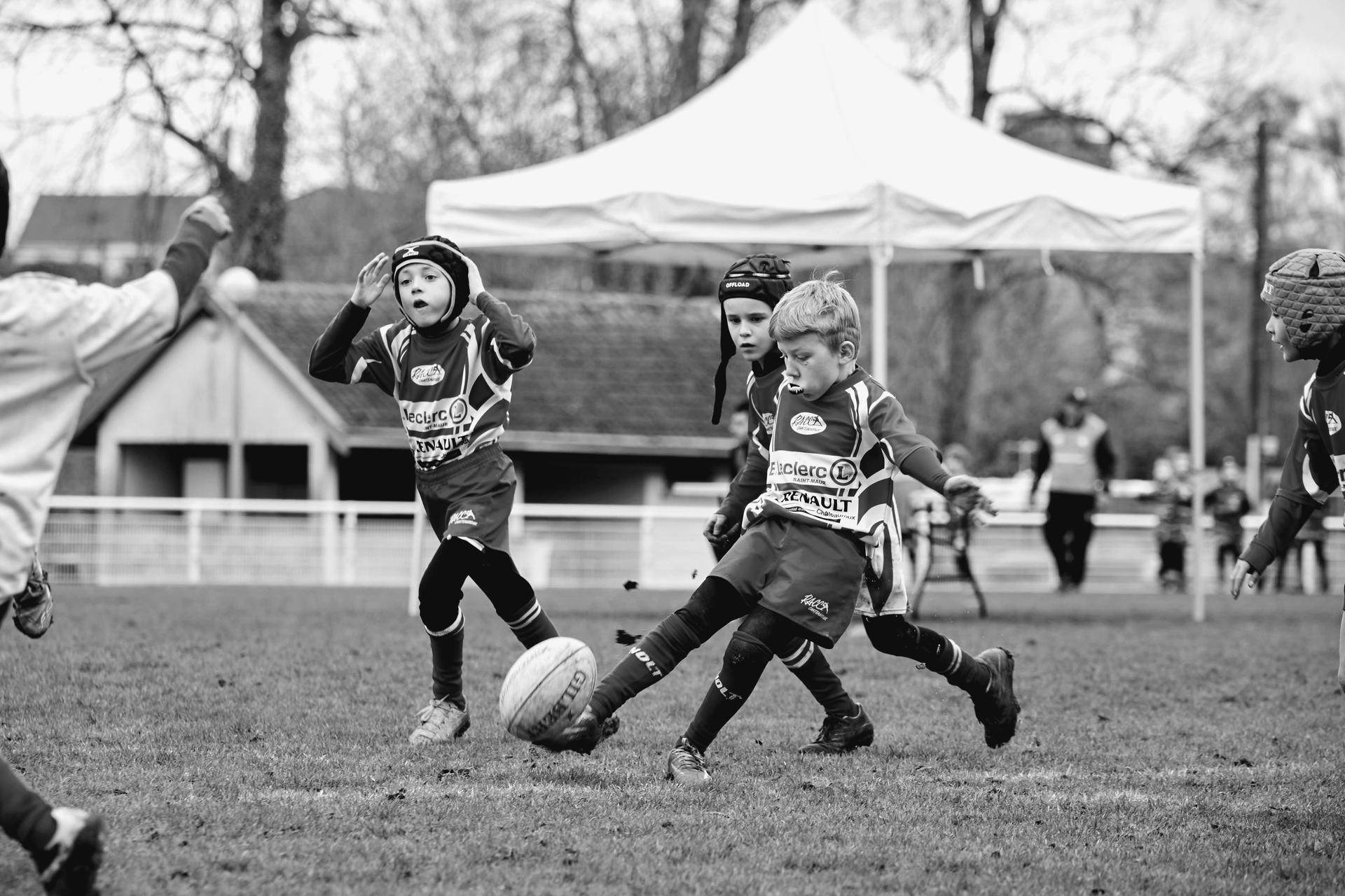 Jeunes joueurs de rugby en plein match, l'un d'eux dégageant le ballon au pied. Photographie en noir et blanc.